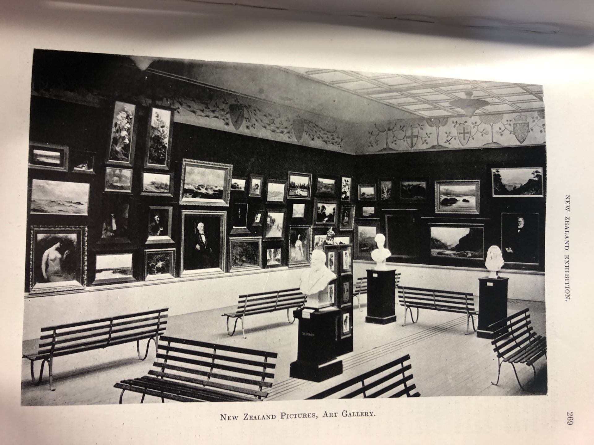 Black and white photo of the New Zealand art gallery at the 1906 International Exhibition in Christchurch. The photo shows two walls covered in 40 paintings, with bench seats in the middle of the room. Some of the paintings are hung 3-4 deep.