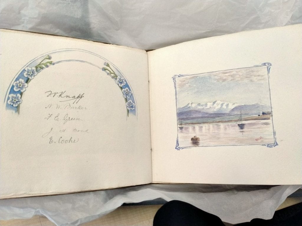 The Suter Gift Book open with a page of signatures and a floral motif on the left, and a watercolour landscape sketch on the right, with mountains and a body of water